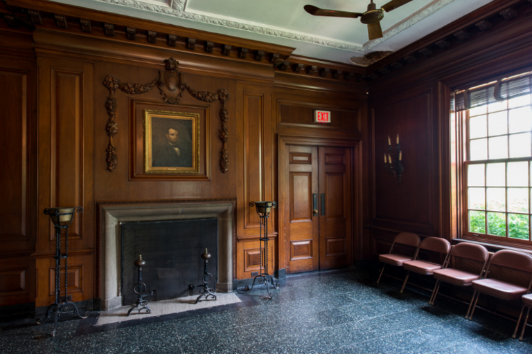 Ornate wood paneled room with fireplace, portrait, and chairs.